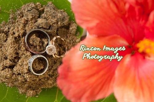 Three wedding rings sitting in sand with a tropical hibiscus flower. Photograph by Rincon Images photography, wedding photographer Rincon Puerto Rico.